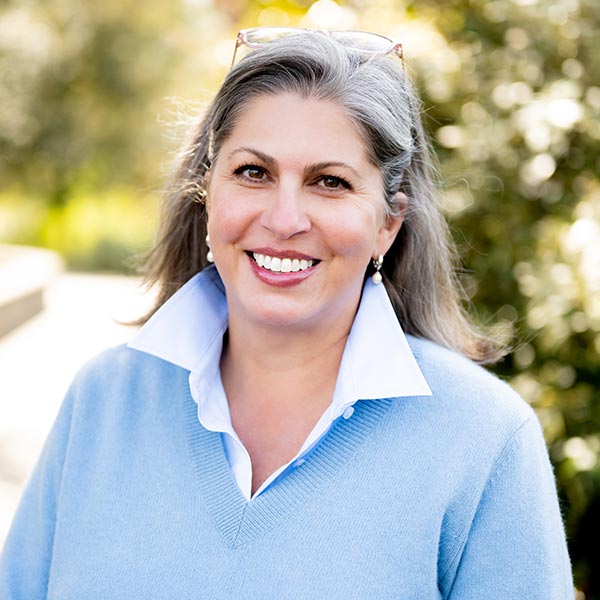 Woman with blonde hair wearing a light blue shirt outdoors with blurred greenery in the background.