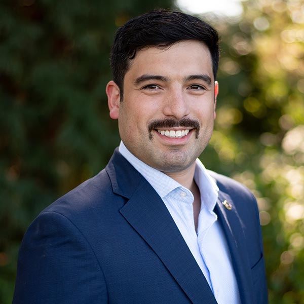 Professional man in dark suit and tie smiling outdoors with green foliage background