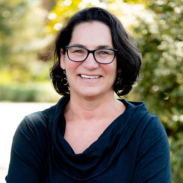 Woman with glasses and dark hair wearing a black top smiling outdoors with blurred foliage in the background.